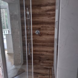 Michał Gwizdek - Kabina prysznicowa z przesuwnymi drzwiami, wykończona płytkami imitującymi drewno i marmur, widoczny odpływ liniowy w podłodze.