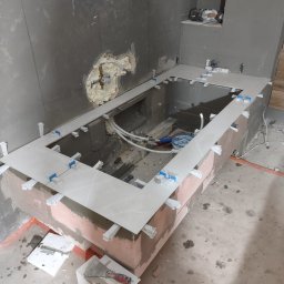 Michał Gwizdek - Montaż płytek na obudowie wanny w łazience, widoczne białe płytki z niebieskimi klinami, instalacja wodna i zaprawa murarska, ściany w szarym tynku.