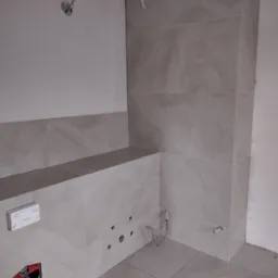 Wnętrze łazienki w trakcie wykańczania: ułożone płytki imitujące marmur na ścianach i podłodze, widoczne otwory na instalacje sanitarne, niezabudowana spłuczka WC, oraz wystające przewody elektryczne.