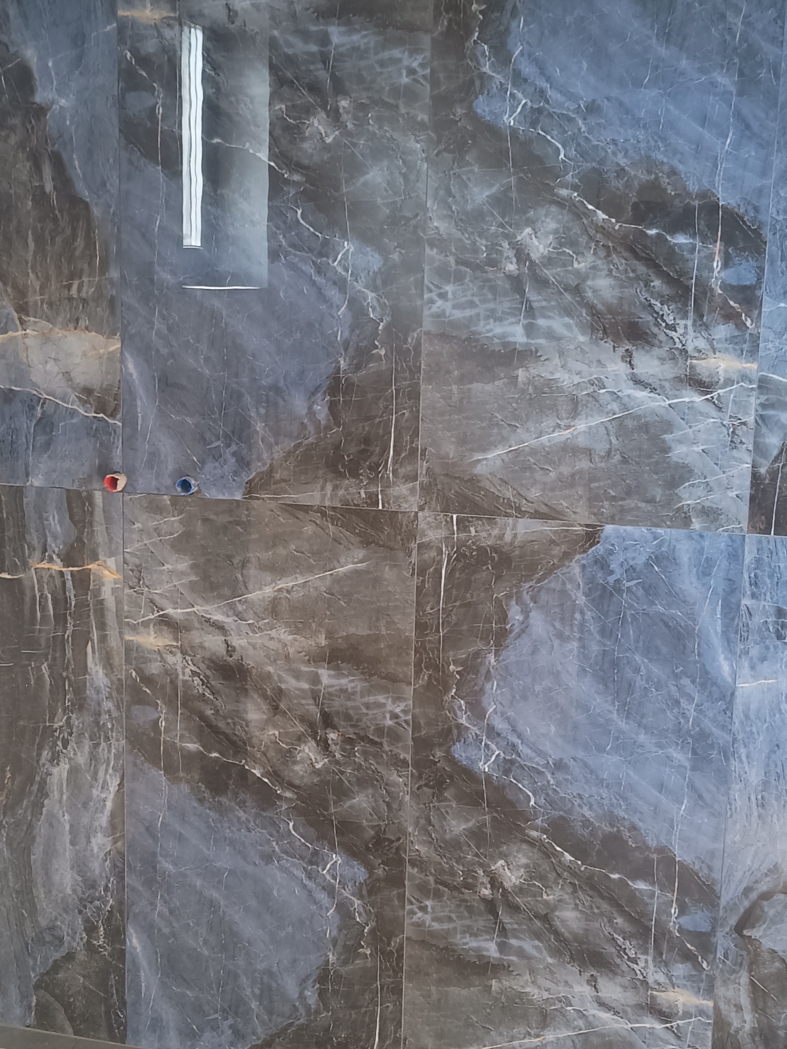 Ściana wyłożona płytkami imitującymi marmur w odcieniach szarości i błękitu, widoczne otwory na instalację wodną i otwór pod odpływ.