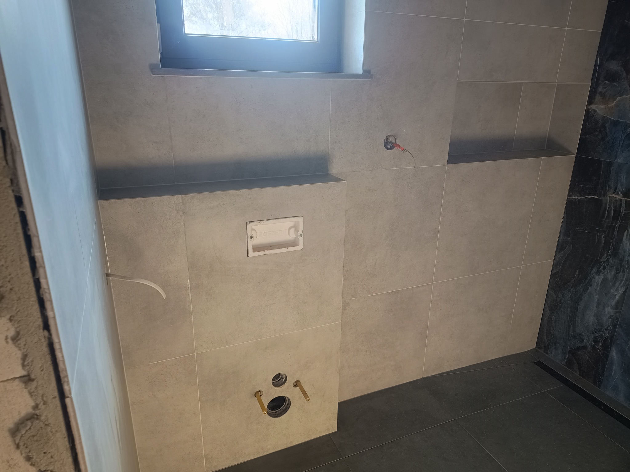 Świeżo wykafelkowana ściana w łazience z widoczną instalacją podtynkową, otworem na spłuczkę i wystającymi rurami do armatury, z fragmentem ściany wykończonej marmurem.