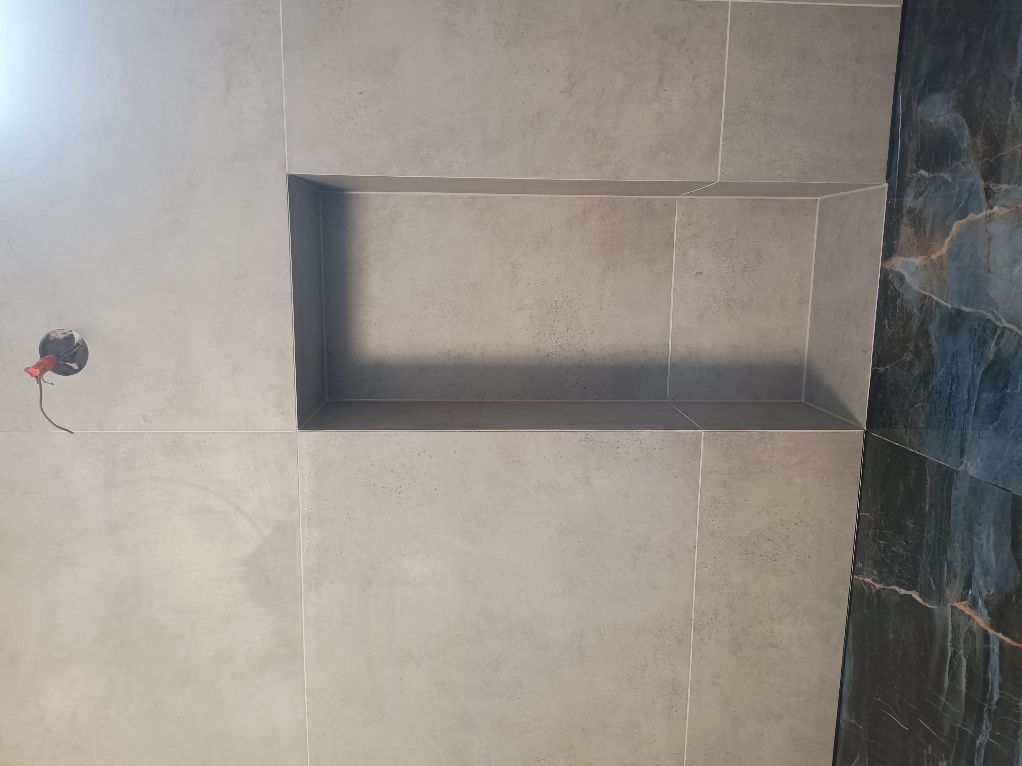 Ściana wyłożona szarymi płytkami z wnęką, obok fragment ściany z płytek imitujących marmur, widoczna puszka elektryczna z wystającymi przewodami.