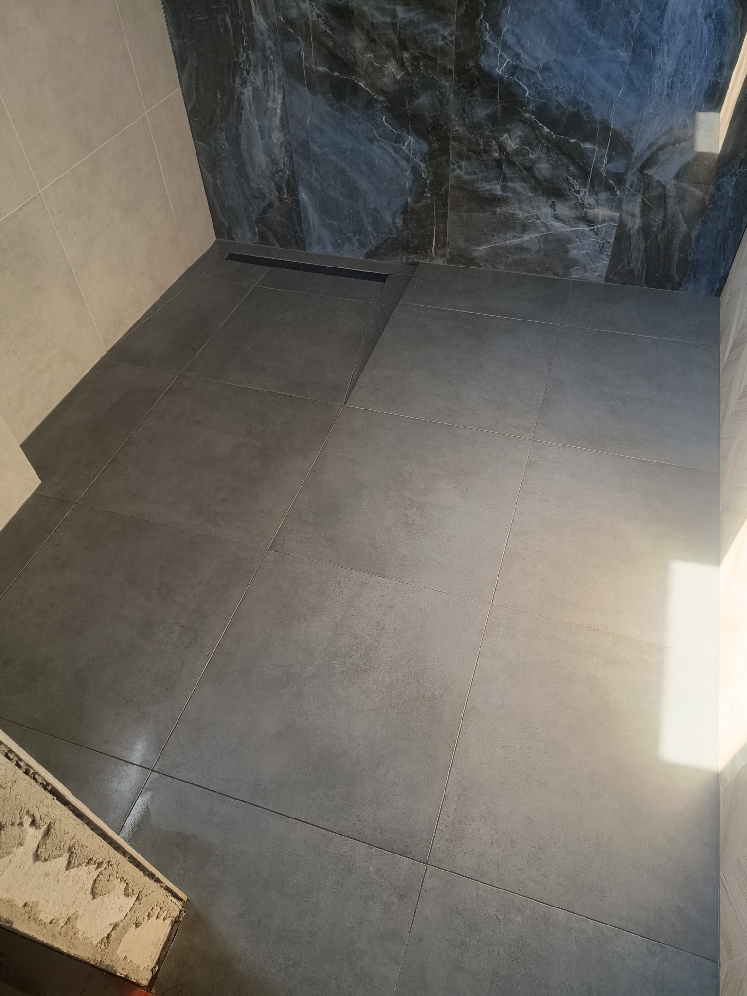 Ukończona łazienka z szarymi płytkami podłogowymi i marmurową ścianą, widoczny odpływ liniowy.