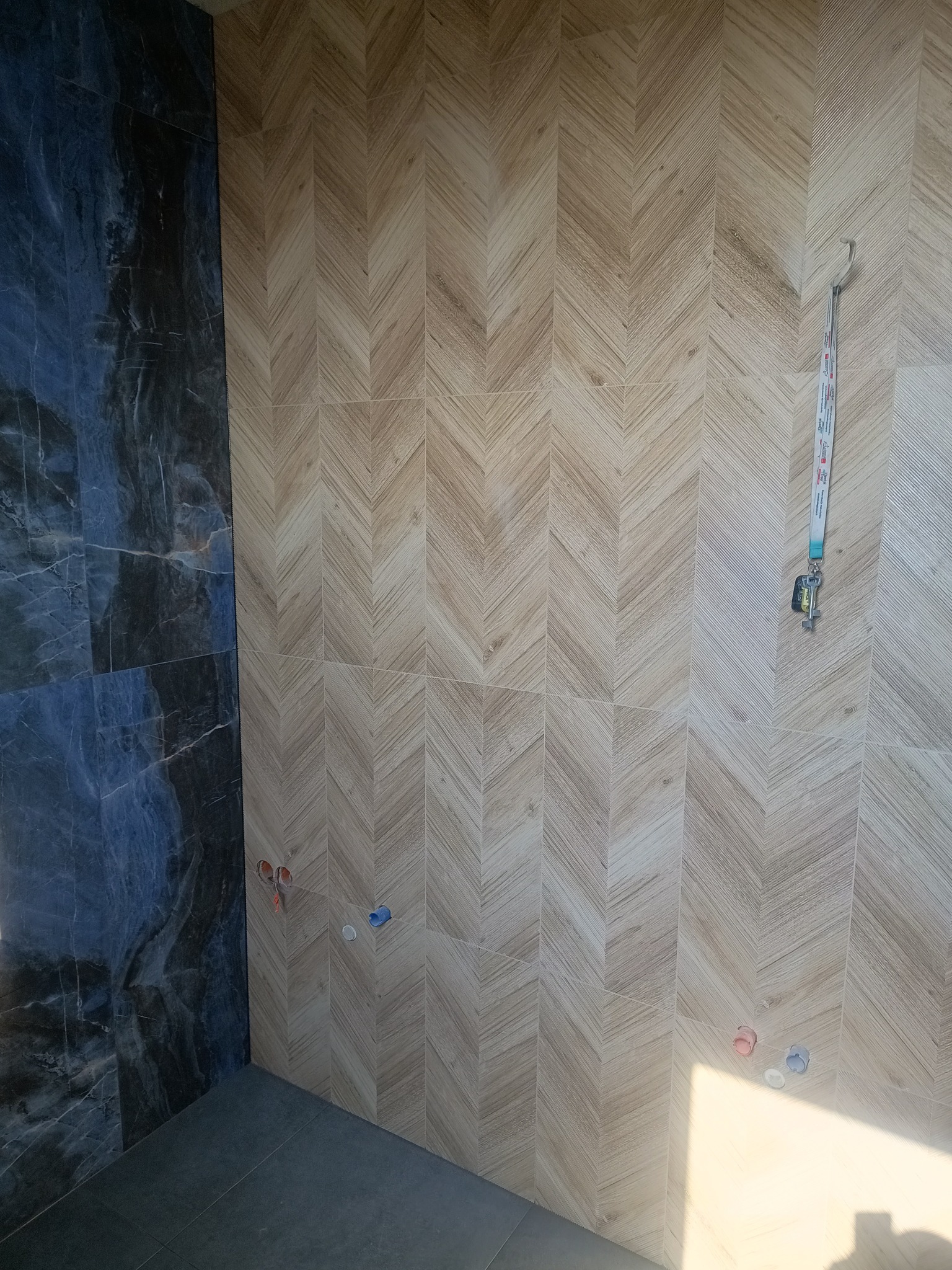 Ściana łazienki wyłożona płytkami imitującymi drewno w jodełkę, kontrastująca z ciemną, marmurkową płytką po lewej stronie. Widoczne wystające rury instalacyjne i smycz z kluczami wisząca na haczyku.