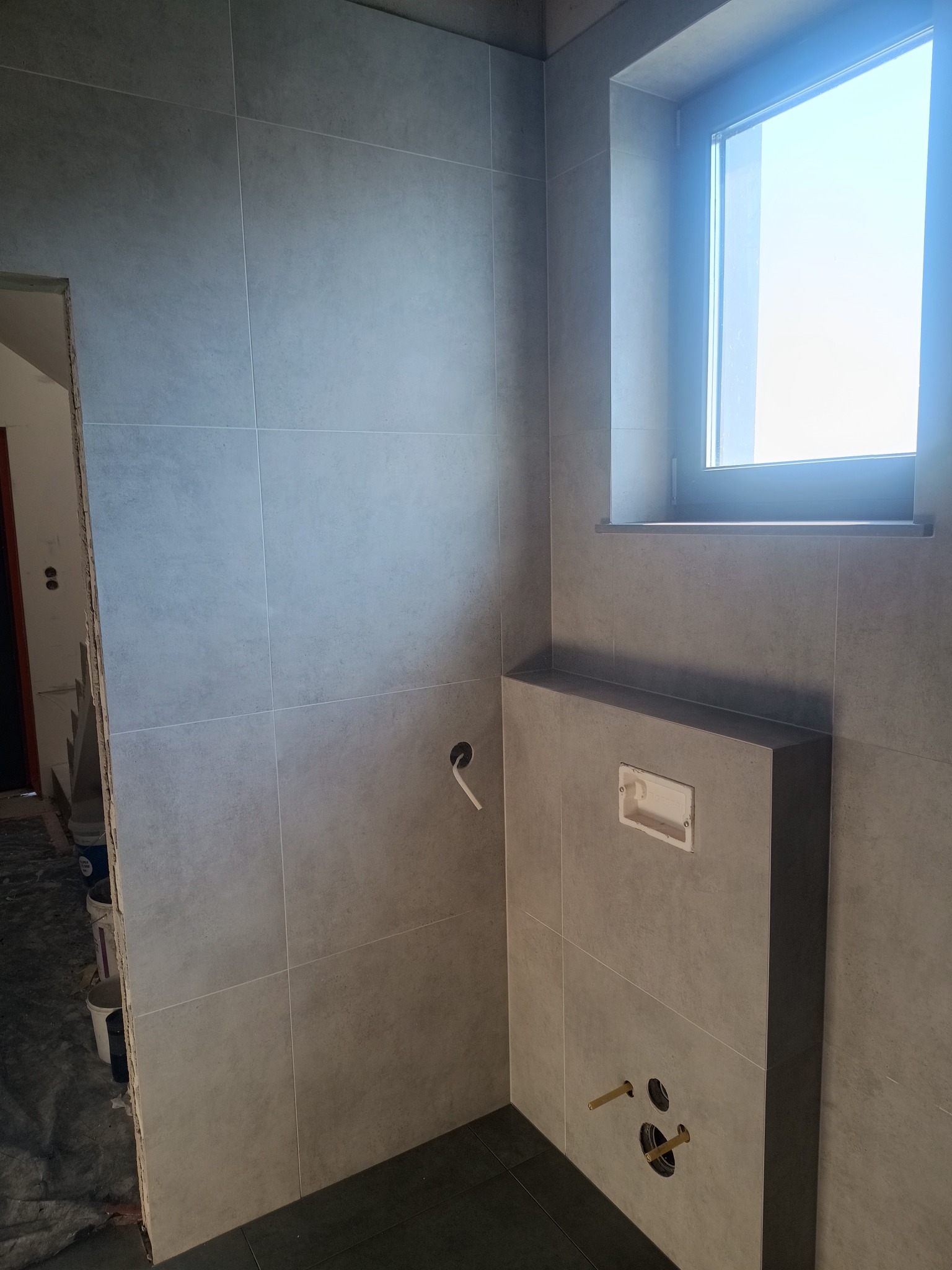 Szare płytki ceramiczne na ścianie w łazience, z widocznym oknem i przygotowanym miejscem na stelaż podtynkowy WC z wystającymi rurami instalacyjnymi.