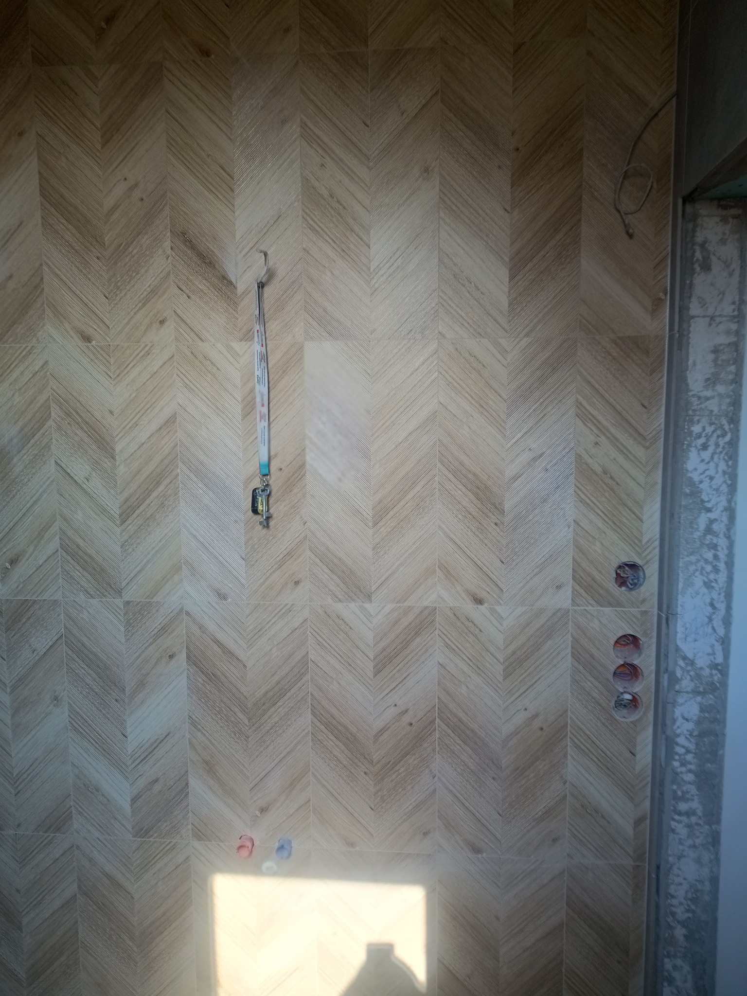 Ściana wyłożona płytkami w jodełkę o drewnopodobnym wzorze; widoczne przyłącza wodne i wystający kabel elektryczny; na haczyku wisi smycz z kluczami.