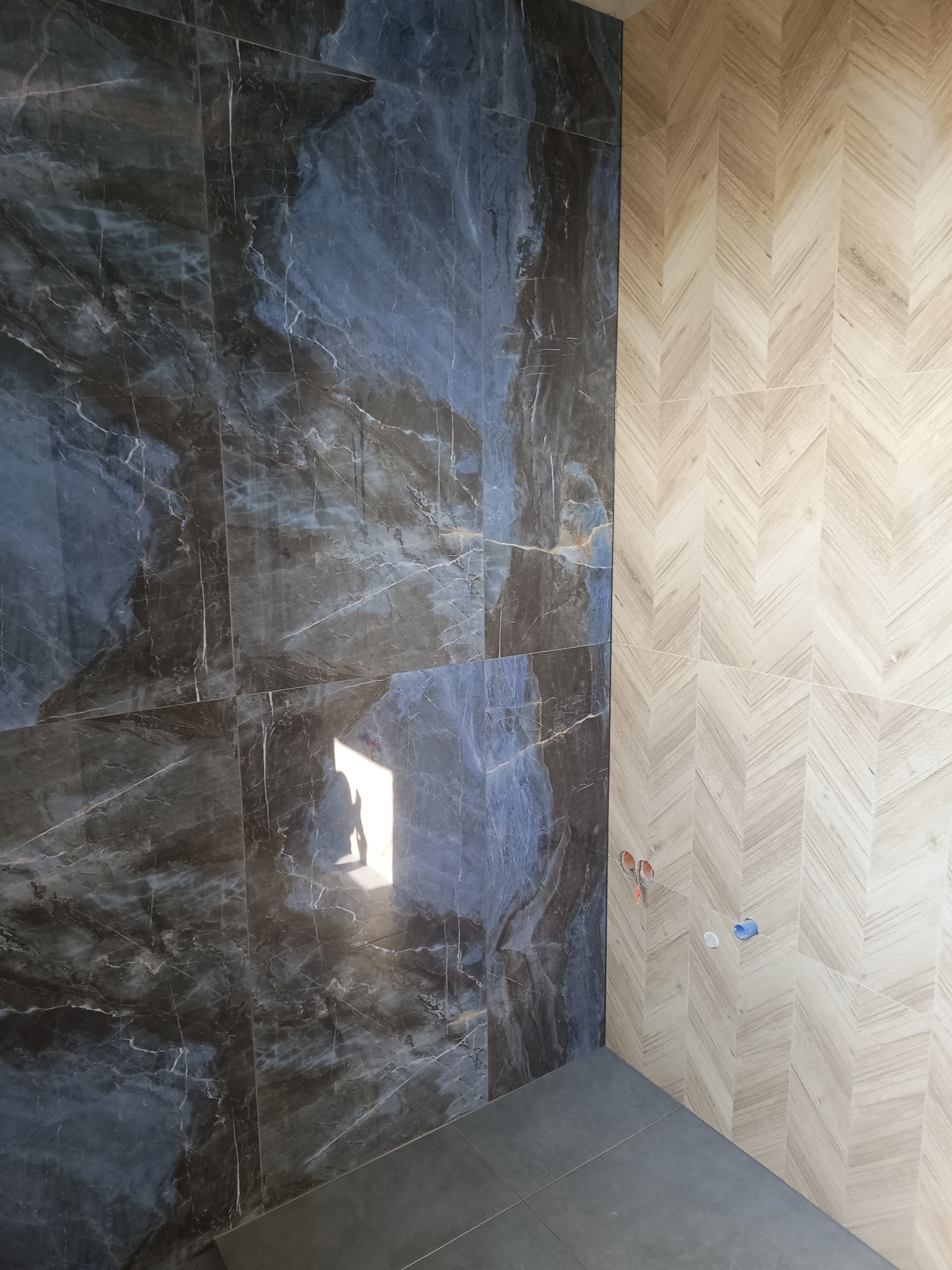 Ukończona ściana w łazience w Ostrowie Wielkopolskim, z połączeniem płytek imitujących marmur w odcieniach szarości i niebieskiego oraz płytek w jasnym odcieniu drewna ułożonych w jodełkę.