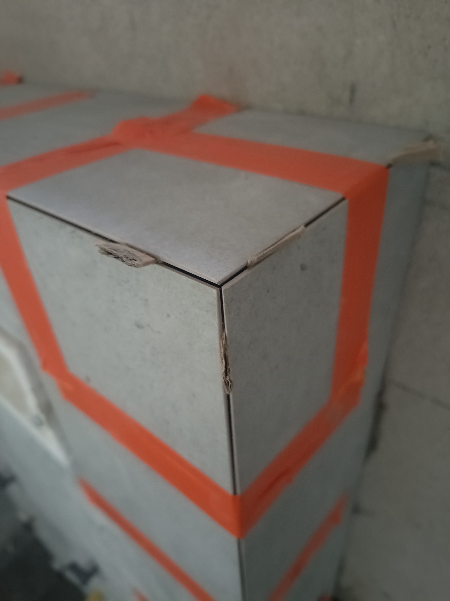 Narożnik obudowy z płytek ceramicznych zabezpieczony pomarańczową taśmą, z widocznym uszkodzeniem krawędzi jednej z płytek.