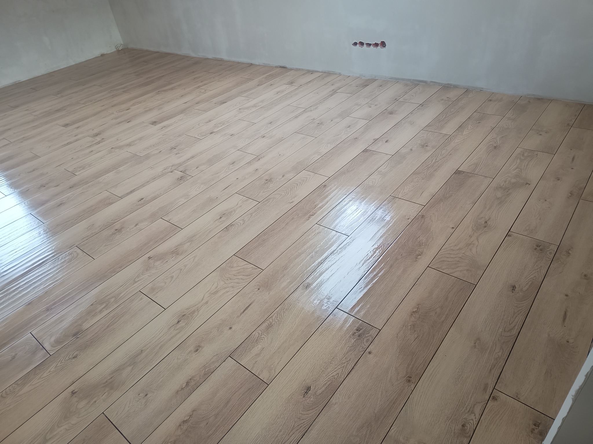 Ułożone panele podłogowe o wzorze jasnego drewna w nowym pomieszczeniu, widoczne gniazdka elektryczne w ścianie, ślady po szpachlowaniu na ścianach.