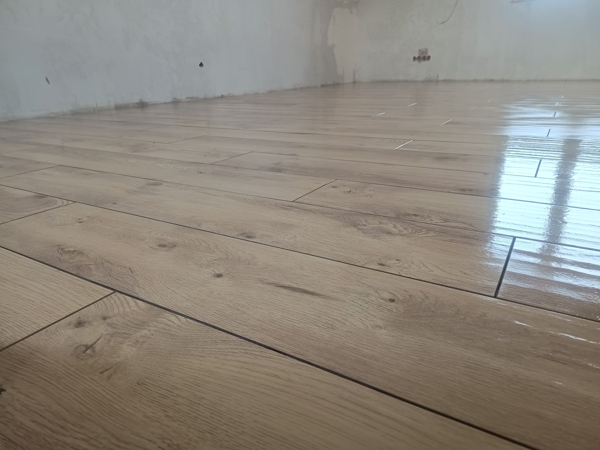 Świeżo ułożone, błyszczące panele podłogowe o wzorze drewna w jasnym odcieniu, z widocznymi szczelinami dylatacyjnymi i surową, niepomalowaną ścianą w tle.