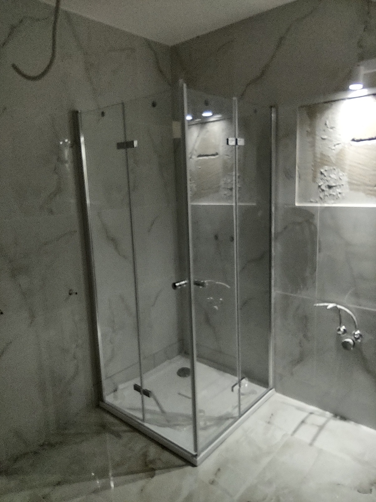 Nowa, narożna kabina prysznicowa z transparentnego szkła, zamontowana w łazience wyłożonej szarymi płytkami imitującymi marmur, widoczny odpływ liniowy w brodziku.