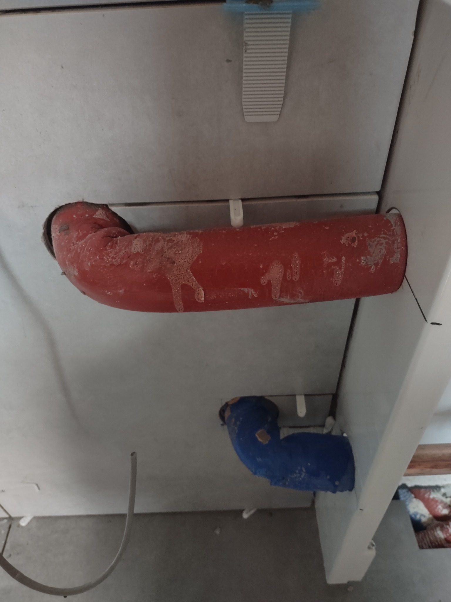 Czerwona rura kanalizacyjna wystająca ze ściany wyłożonej szarymi płytkami, z widocznymi śladami zaprawy, obok niebieska rura kanalizacyjna