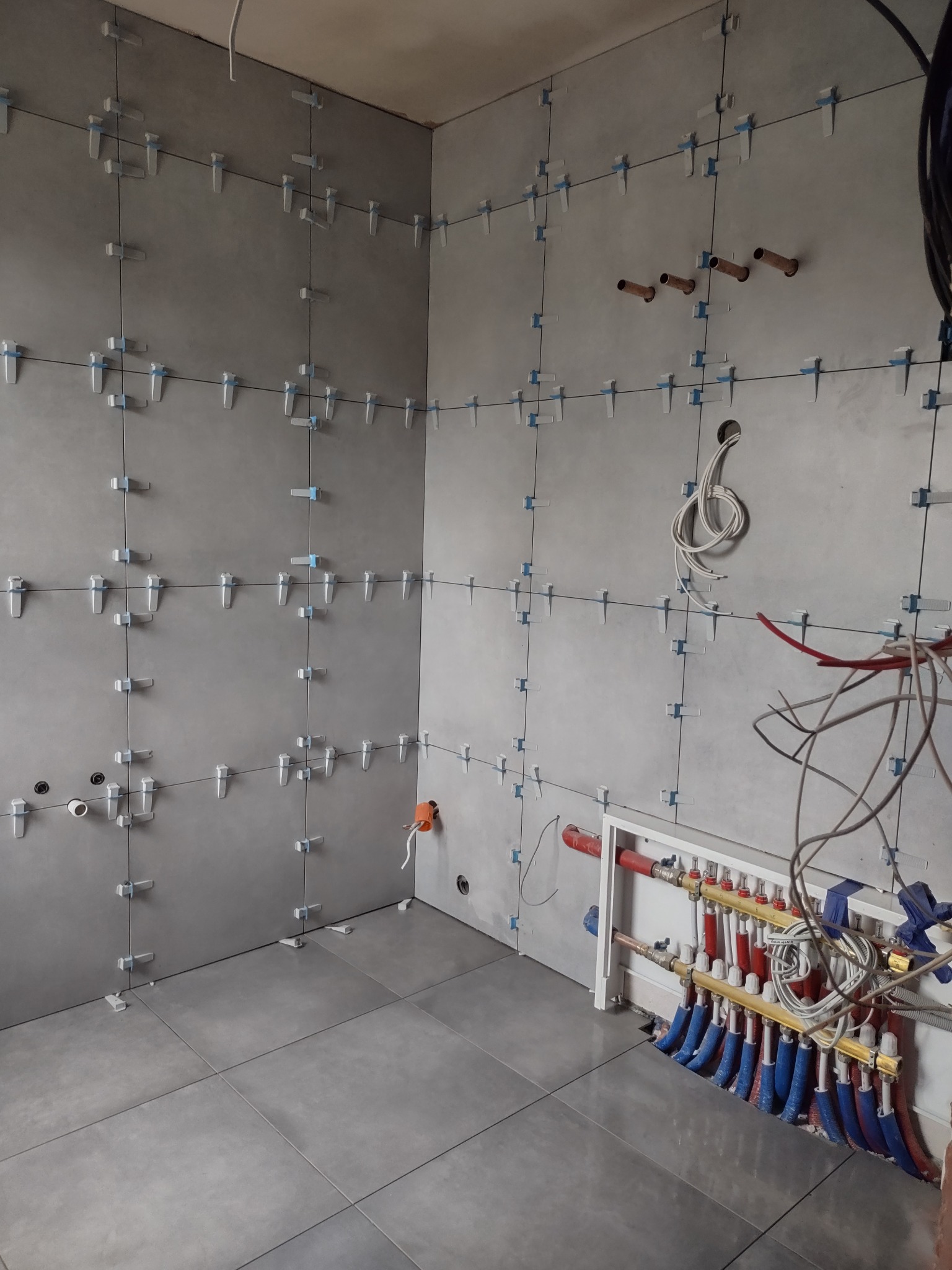 Szare płytki ułożone na ścianach i podłodze z użyciem klipsów poziomujących, widoczna instalacja wodna i elektryczna w trakcie prac wykończeniowych.