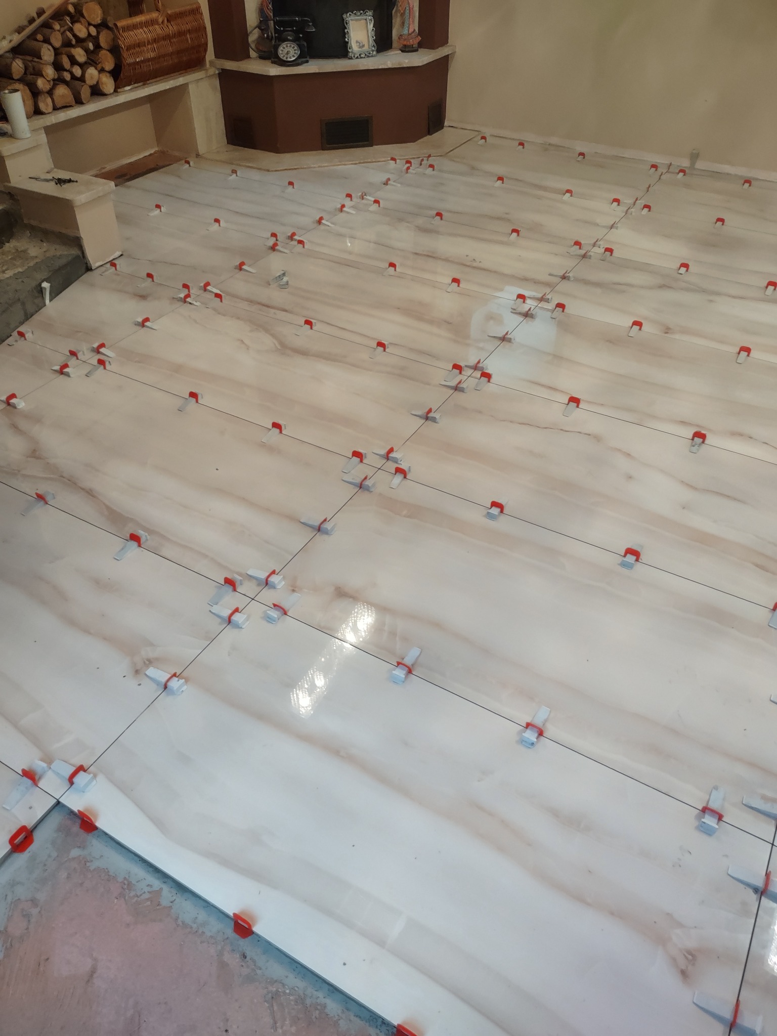 Podłoga z jasnych, marmurkowych płytek, w trakcie układania, z użyciem systemu poziomowania płytek (klipsy i kliny), widoczny fragment ściany z boazerią i kominkiem w tle.