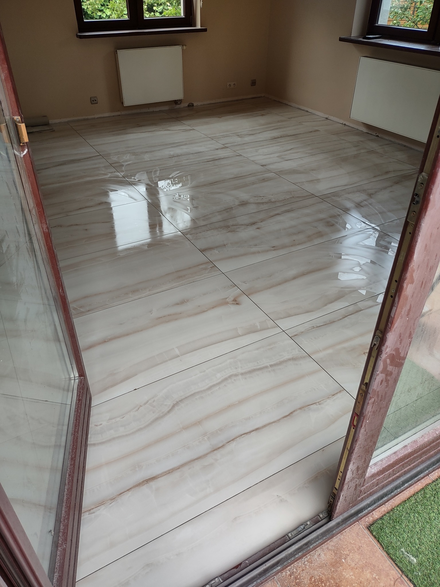 Jasne, błyszczące płytki podłogowe imitujące marmur, ułożone w nowym, pustym pokoju, widoczne przez otwarte drzwi balkonowe.