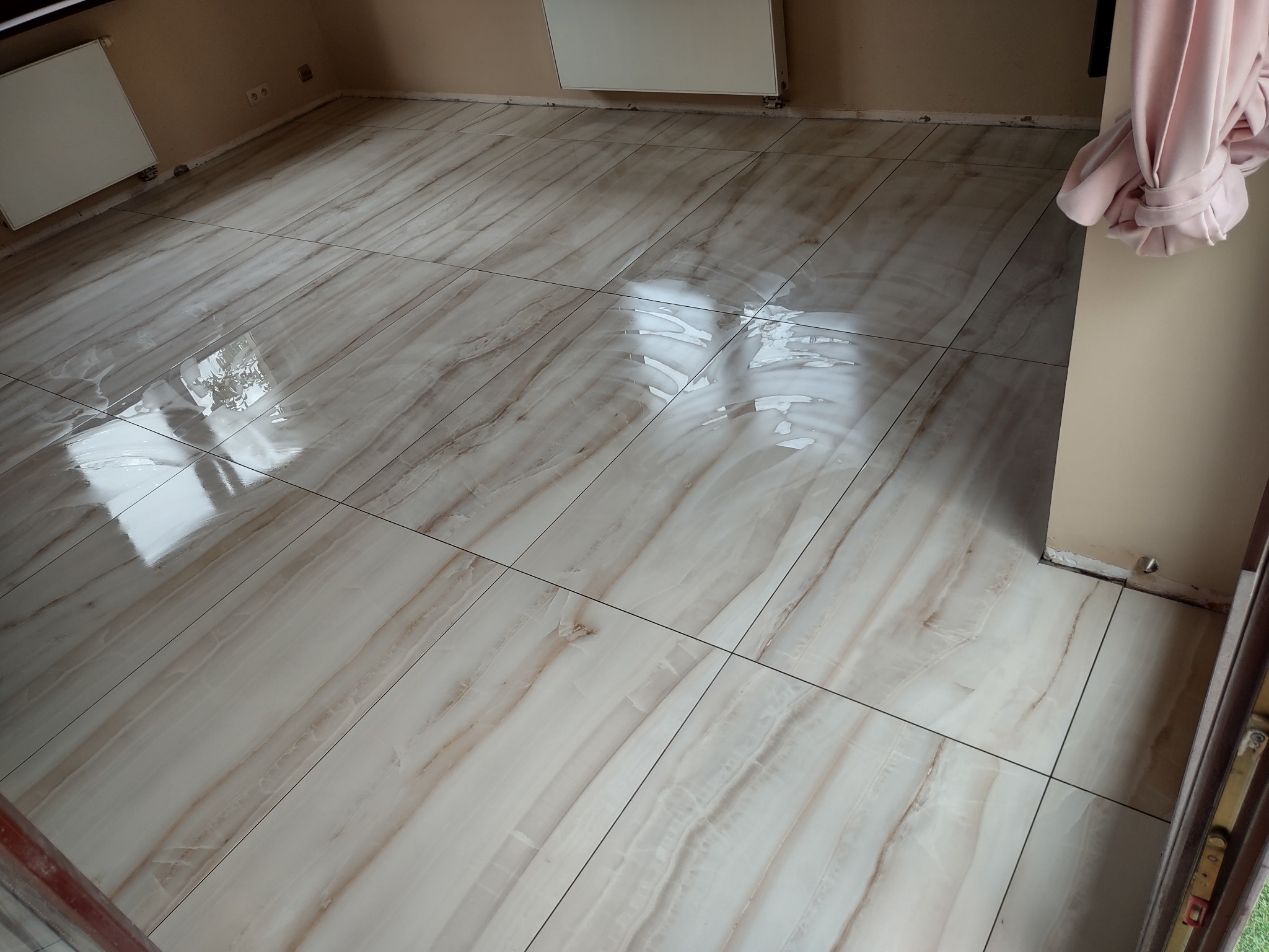 Ułożone, błyszczące płytki podłogowe w odcieniach beżu i brązu z wyraźnym wzorem imitującym marmur, widoczne niedociągnięcia przy ścianach.