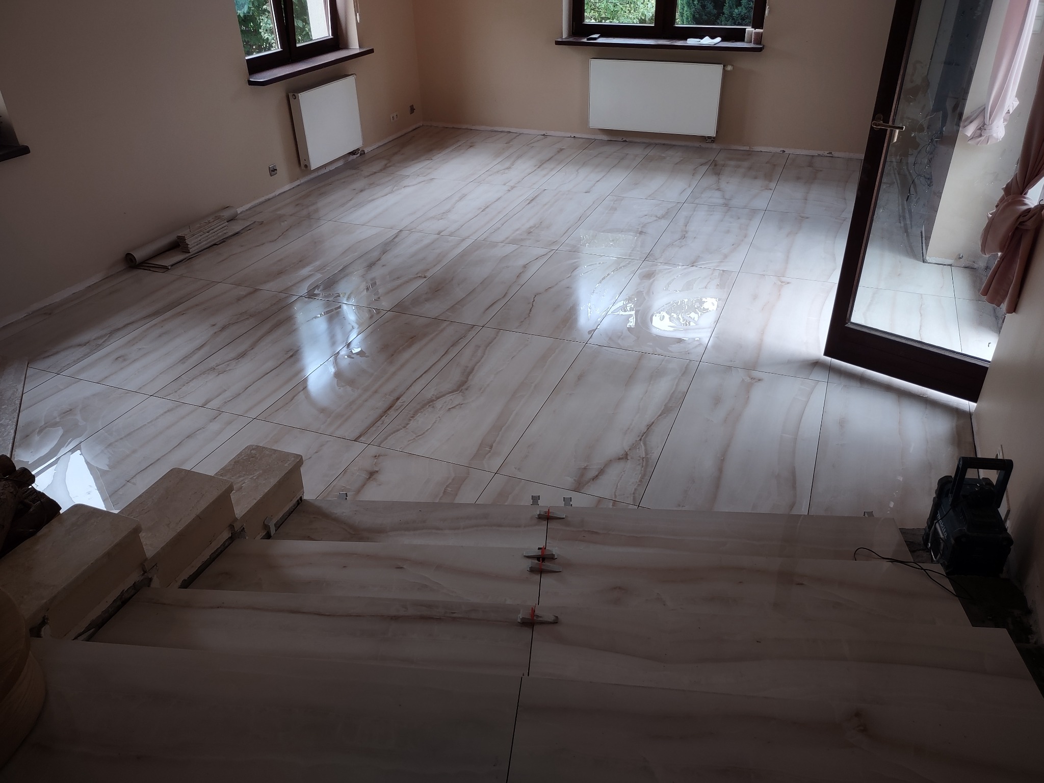 Wykończone schody i podłoga z płytek imitujących marmur, z widocznymi krzyżykami dystansowymi i odkurzaczem budowlanym w rogu pomieszczenia.