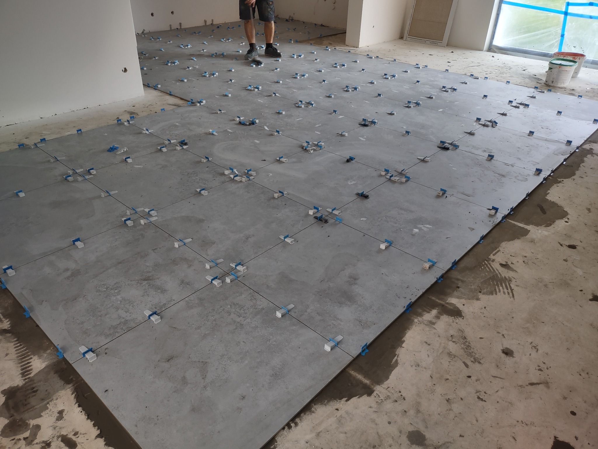 Układanie szarych płytek podłogowych z użyciem systemu poziomowania, widoczne krzyżyki dystansowe i kliny, fragment podłogi niepokryty płytkami.