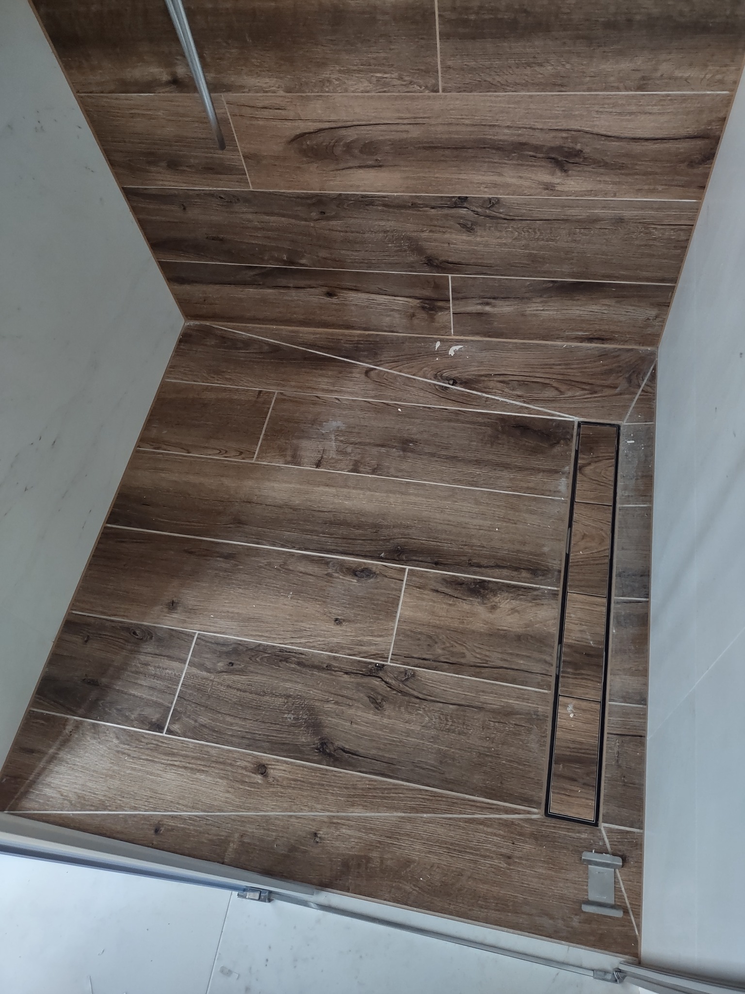 Wykończona kabina prysznicowa z brązowymi płytkami imitującymi drewno i liniowym odpływem, widoczne białe ściany i fragment kabiny.