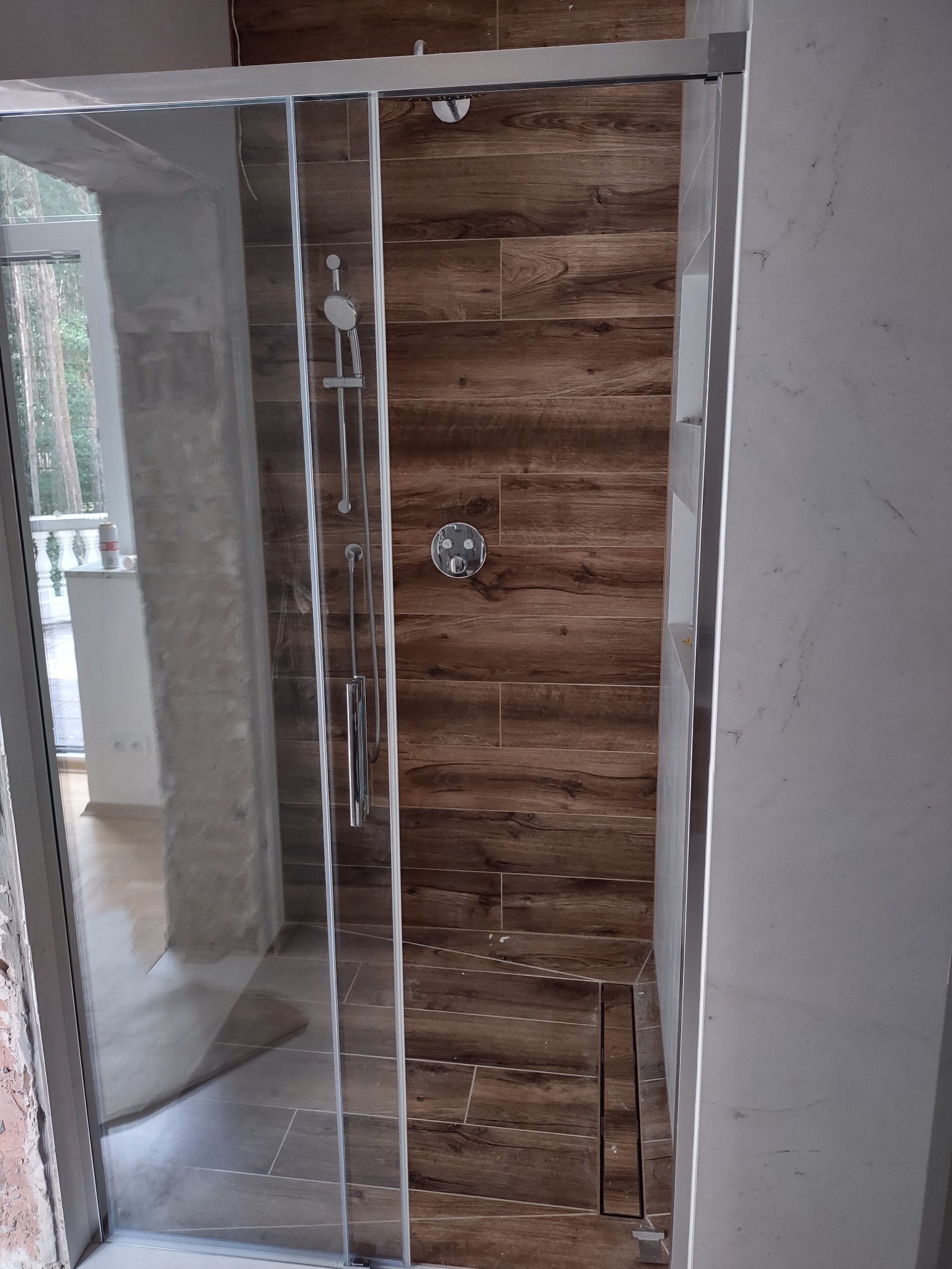 Kabina prysznicowa z przesuwnymi drzwiami, wykończona płytkami imitującymi drewno i marmur, widoczny odpływ liniowy w podłodze.