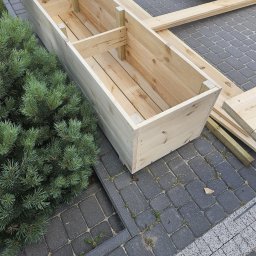 TOMASZ BARCIKOWSKI - Surowa, drewniana skrzynia na rośliny z wewnętrzną przegrodą, ustawiona na kostce brukowej obok iglastego krzewu, z widocznymi deskami i niebieską plandeką w tle.