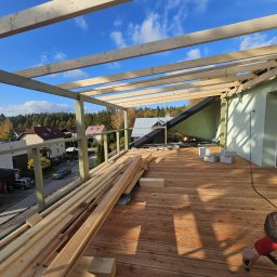 TOMASZ BARCIKOWSKI - Montaż zadaszenia tarasu drewnianego z widokiem na okolicę, deski tarasowe ułożone, konstrukcja z belek w trakcie budowy, narzędzia i materiały budowlane na tarasie.