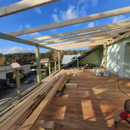 Montaż zadaszenia tarasu drewnianego z widokiem na okolicę, deski tarasowe ułożone, konstrukcja z belek w trakcie budowy, narzędzia i materiały budowlane na tarasie.