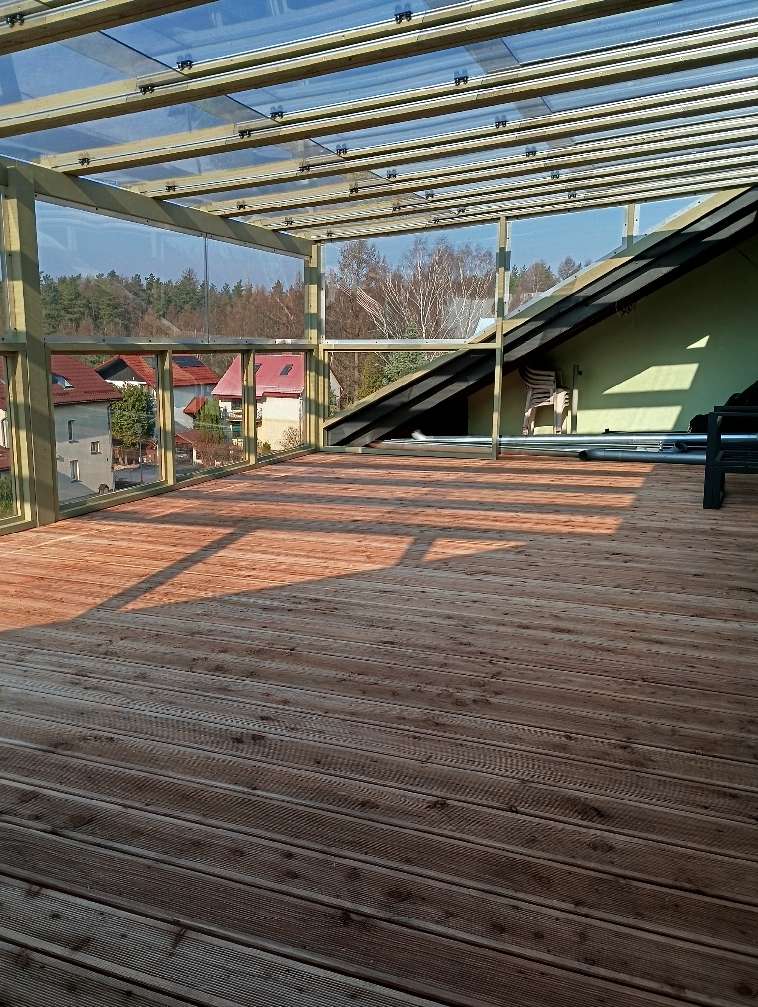 Zadaszony taras z drewnianą podłogą i przeszklonym dachem, widok na okoliczne domy i drzewa.