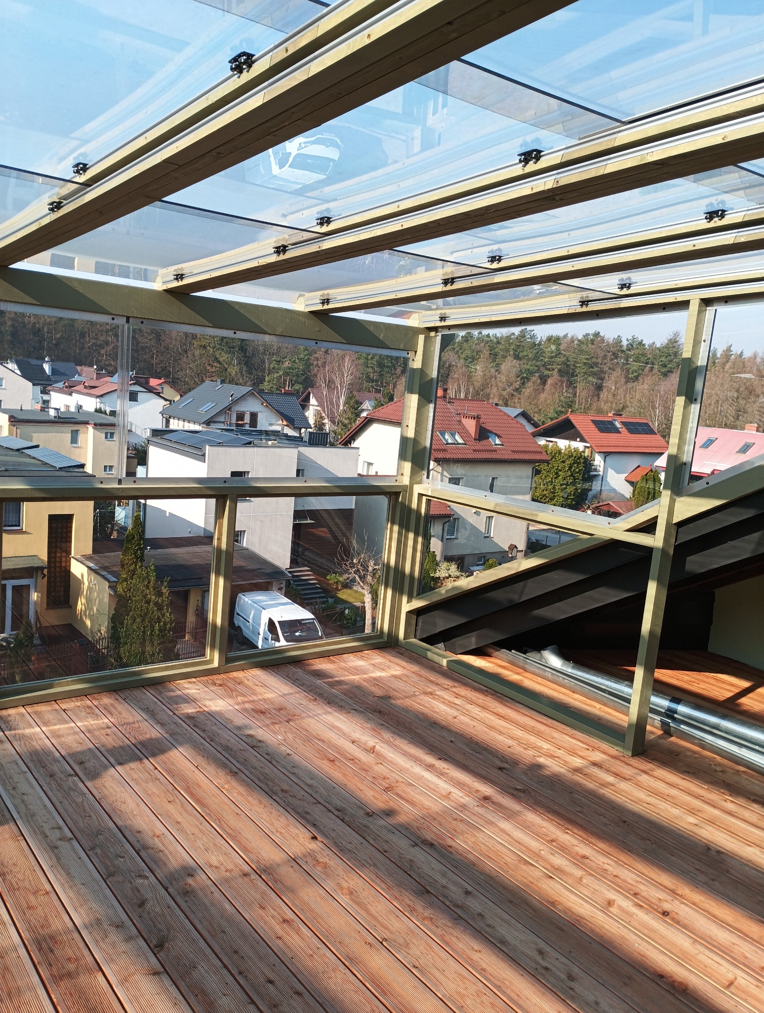 Zadaszony taras z drewnianą podłogą i szklanymi ścianami, widok na zabudowę jednorodzinną i las w tle.