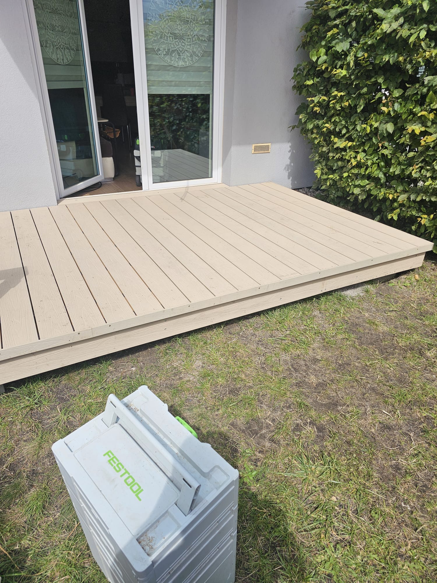 Beżowy taras z desek kompozytowych przylegający do domu z otwartymi drzwiami balkonowymi, obok zielony żywopłot, na trawie skrzynka narzędziowa marki Festool.