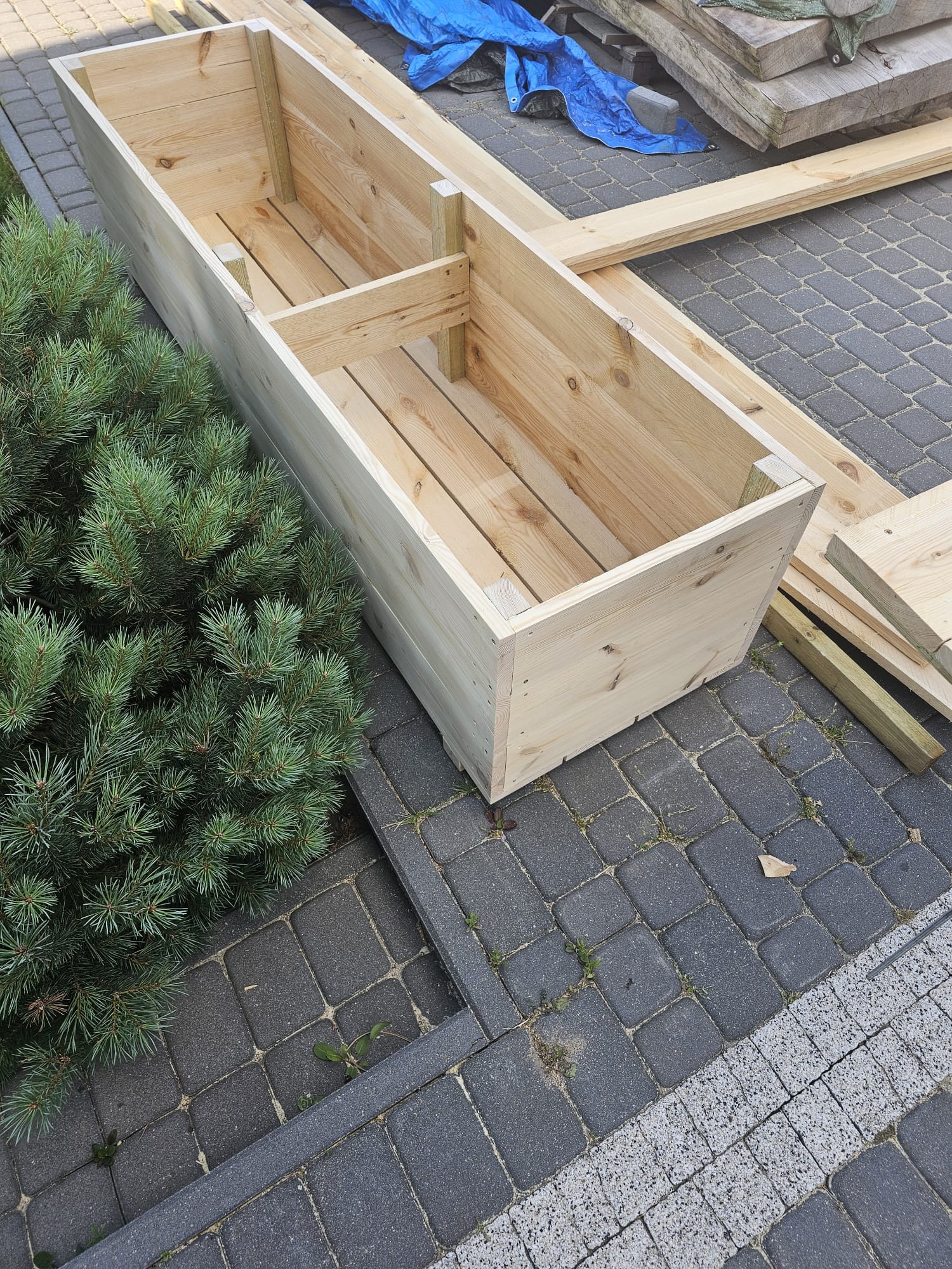 Surowa, drewniana skrzynia na rośliny z wewnętrzną przegrodą, ustawiona na kostce brukowej obok iglastego krzewu, z widocznymi deskami i niebieską plandeką w tle.