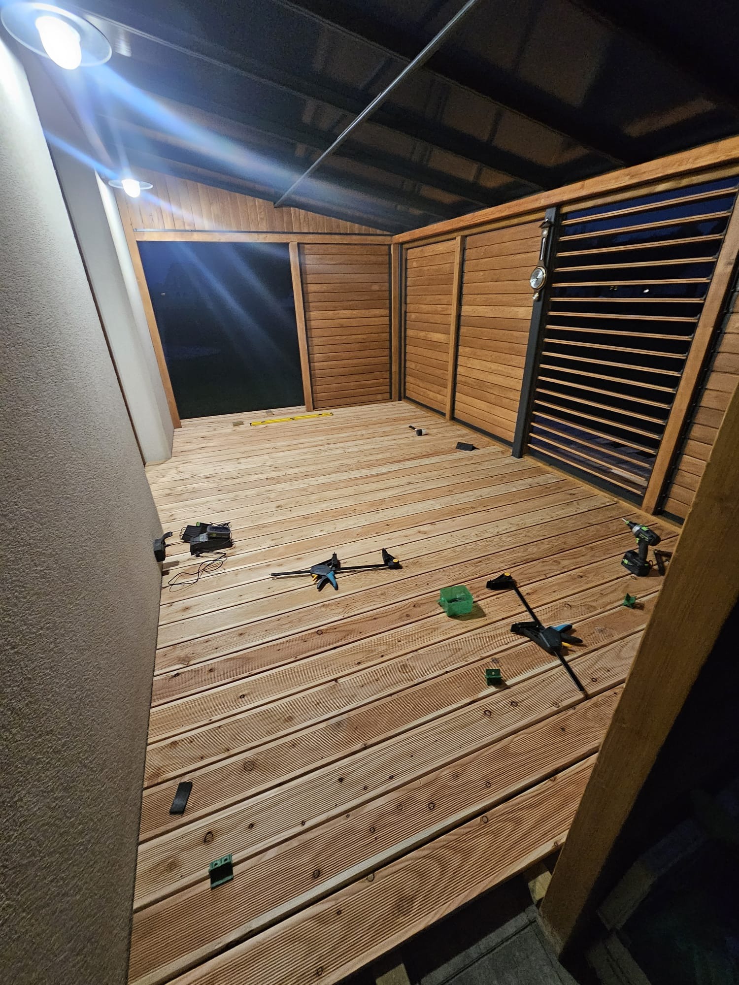 Montaż tarasu z desek na legarach w zadaszonej konstrukcji, widoczne narzędzia i poziomica, oświetlenie górne.