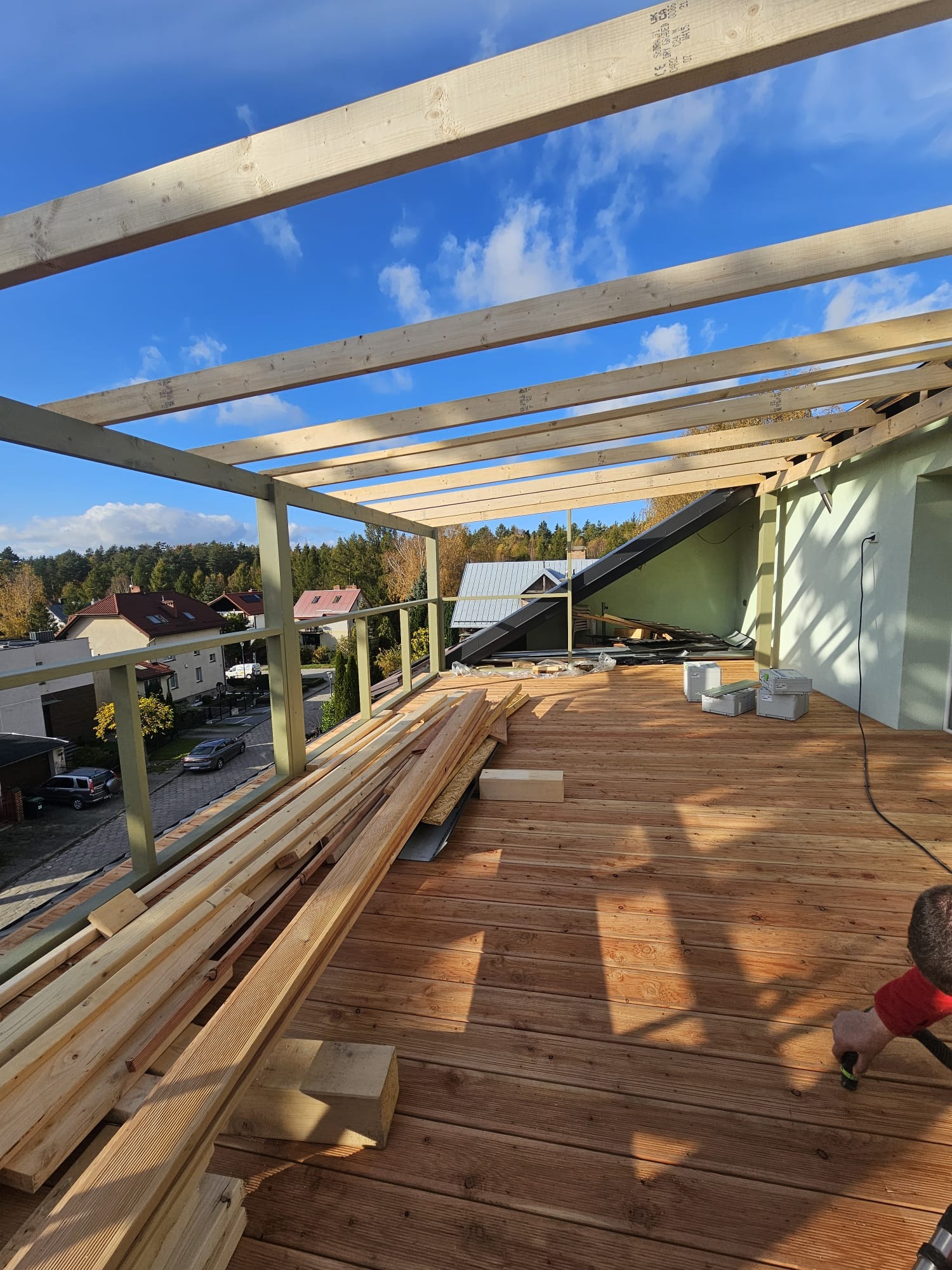 Montaż zadaszenia tarasu drewnianego z widokiem na okolicę, deski tarasowe ułożone, konstrukcja z belek w trakcie budowy, narzędzia i materiały budowlane na tarasie.