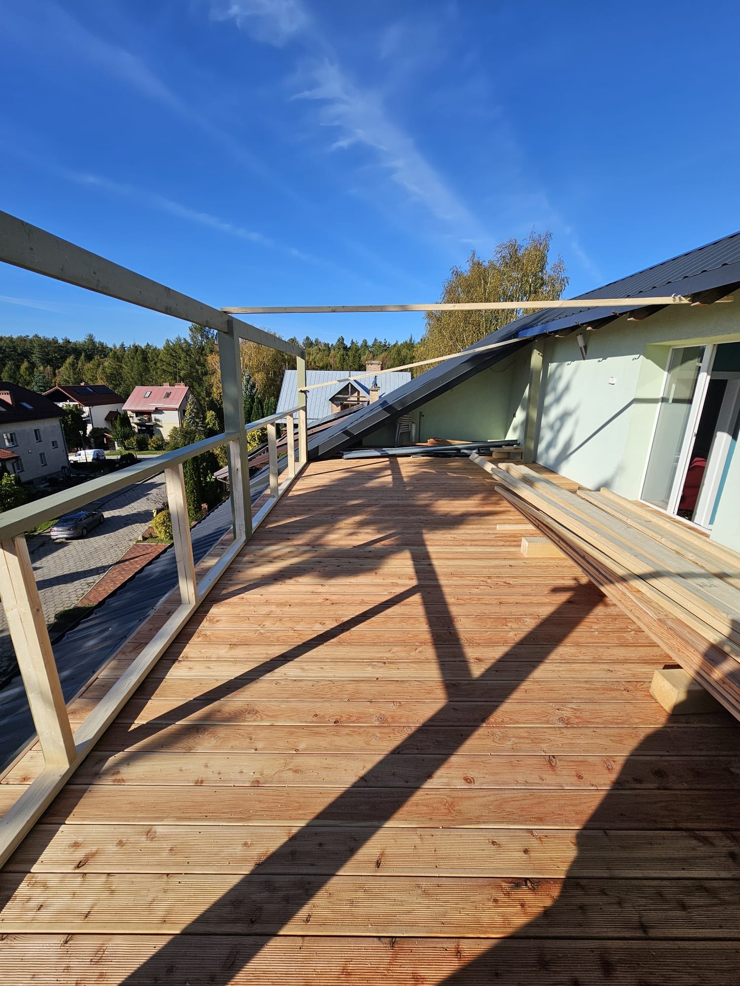 Budowa tarasu na dachu domu jednorodzinnego w słoneczny dzień. Widoczne deski tarasowe, konstrukcja drewniana balustrady oraz przygotowane elementy do dalszego montażu. W tle zabudowa jednorodzinna...