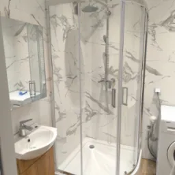 Nowoczesna, jasna łazienka z kabiną prysznicową narożną, umywalką na szafce z drewnopodobnym frontem i pralką Beko, wykończona białymi płytkami imitującymi marmur.