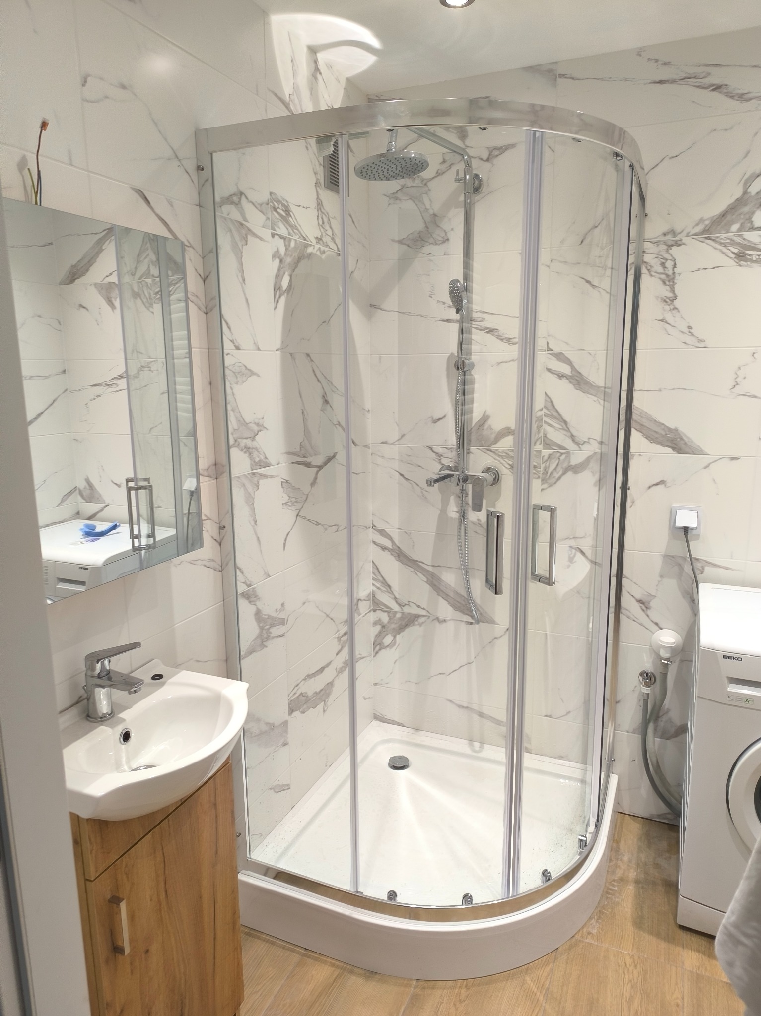 Nowoczesna, jasna łazienka z kabiną prysznicową narożną, umywalką na szafce z drewnopodobnym frontem i pralką Beko, wykończona białymi płytkami imitującymi marmur.