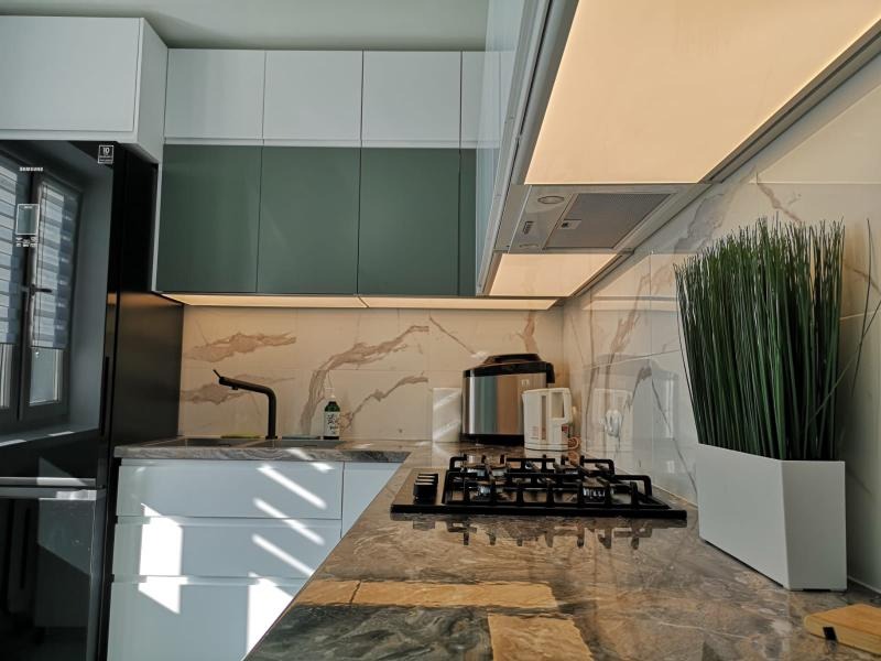 Nowoczesna kuchnia z białymi i zielonymi szafkami, marmurowym blatem, czarną armaturą i płytą gazową oraz dekoracyjną trawą w białej donicy.