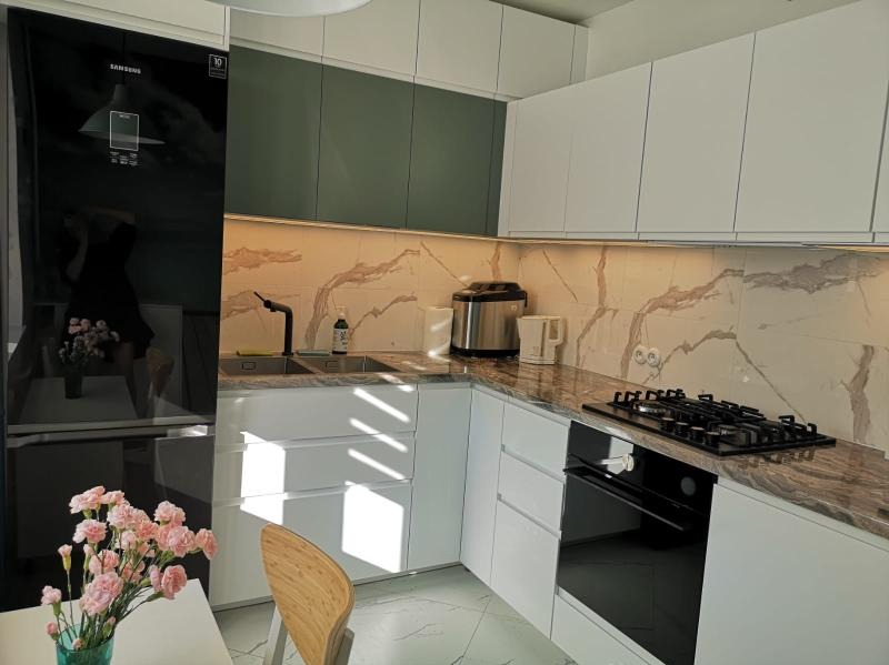 Nowoczesna kuchnia narożna z białymi i oliwkowymi szafkami, marmurowym blatem i backsplashiem, z czarną armaturą i sprzętem AGD, oświetlona listwą LED pod szafkami górnymi.