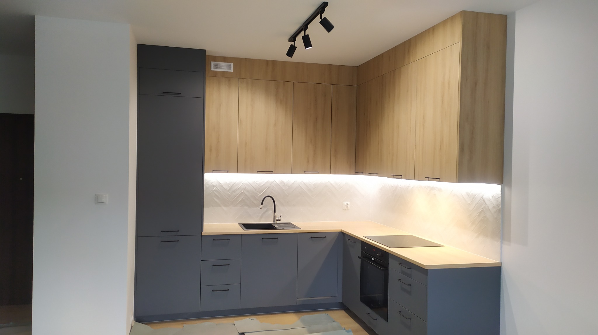 Nowoczesna kuchnia z szarymi dolnymi szafkami i drewnianymi górnymi, oświetlona listwą LED pod szafkami, z czarnym zlewem i baterią na tle białych płytek ułożonych w jodełkę.