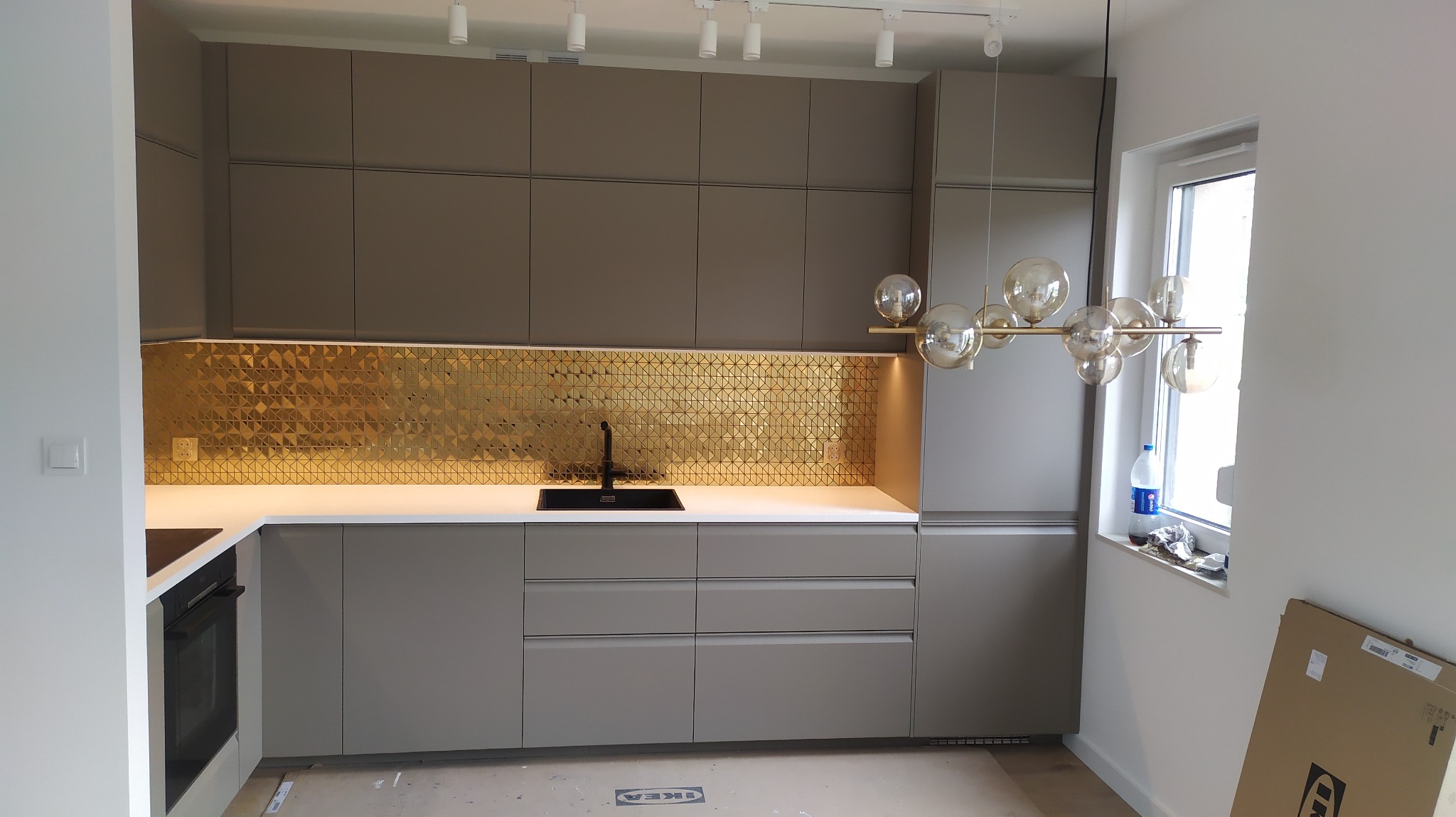 Szara zabudowa kuchenna z złotą mozaiką nad blatem, czarny zlew i bateria, nowoczesna lampa z szklanymi kulami, karton Ikea w rogu pomieszczenia.