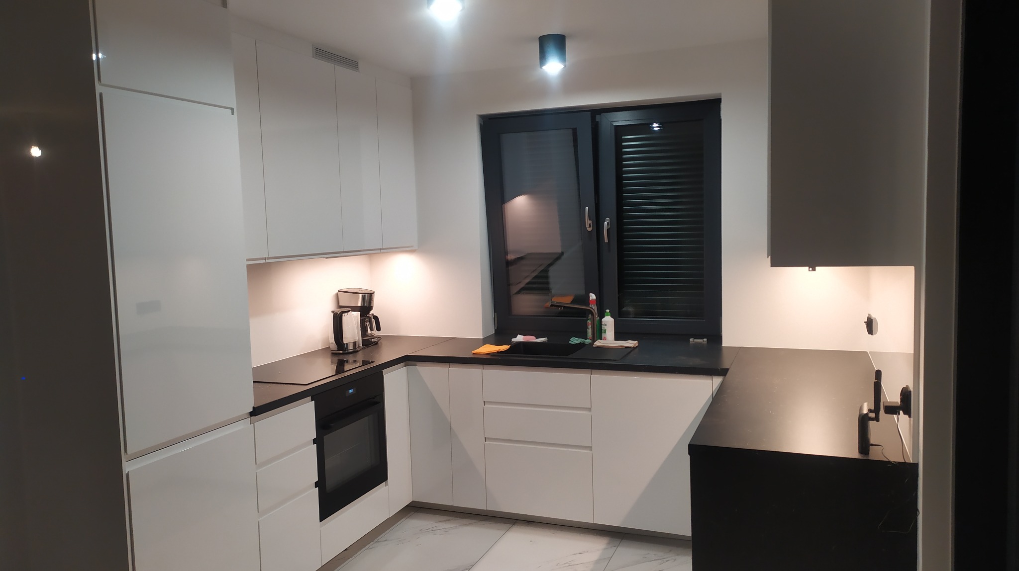 Nowoczesna kuchnia z białymi, lakierowanymi frontami, czarnym blatem i oknem z żaluzjami poziomymi, widoczne oświetlenie podszafkowe i ekspres do kawy.