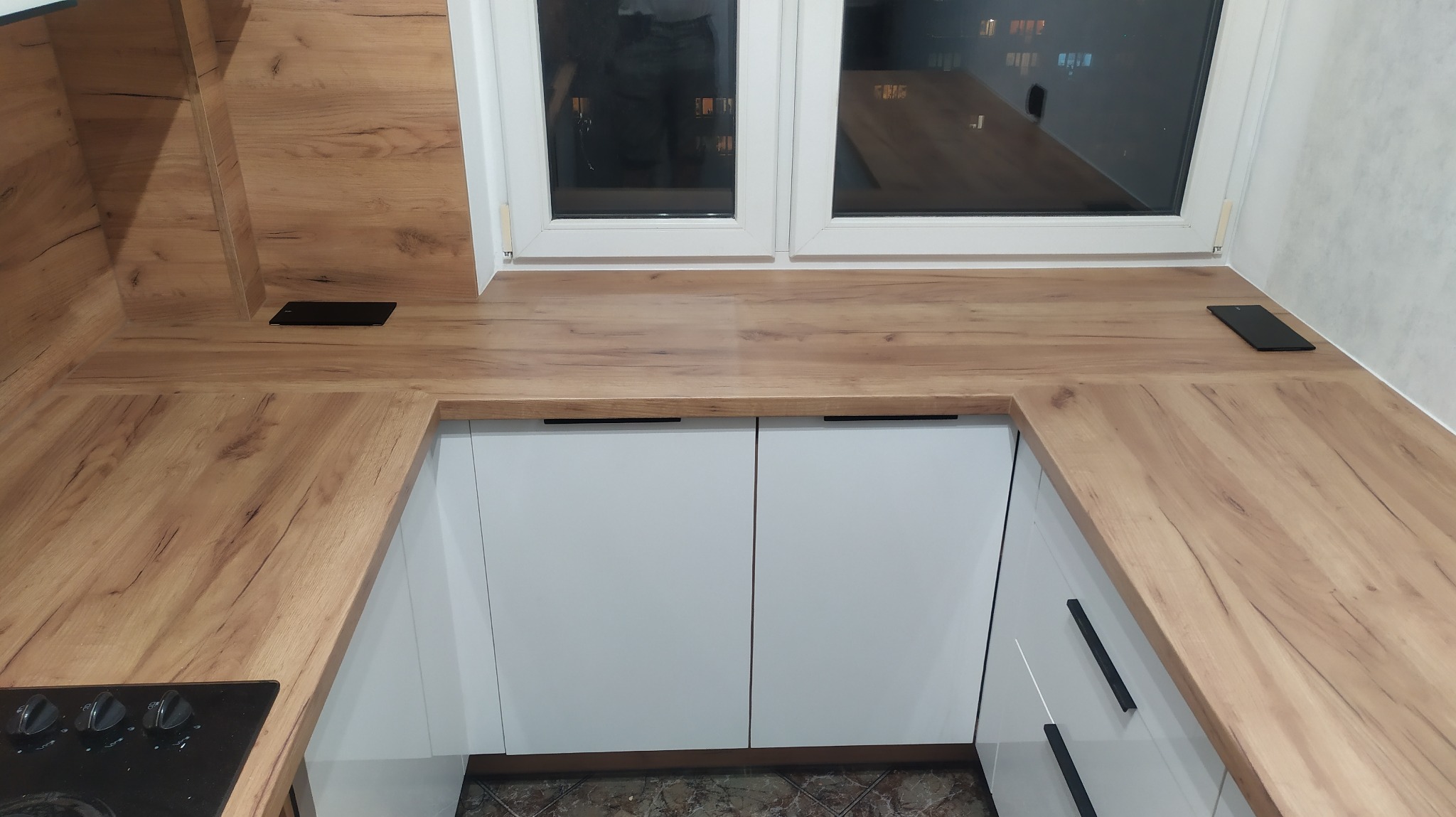 Fragment kuchni z drewnianym blatem, białymi szafkami z czarnymi uchwytami i wbudowanym gniazdkiem elektrycznym przy oknie.