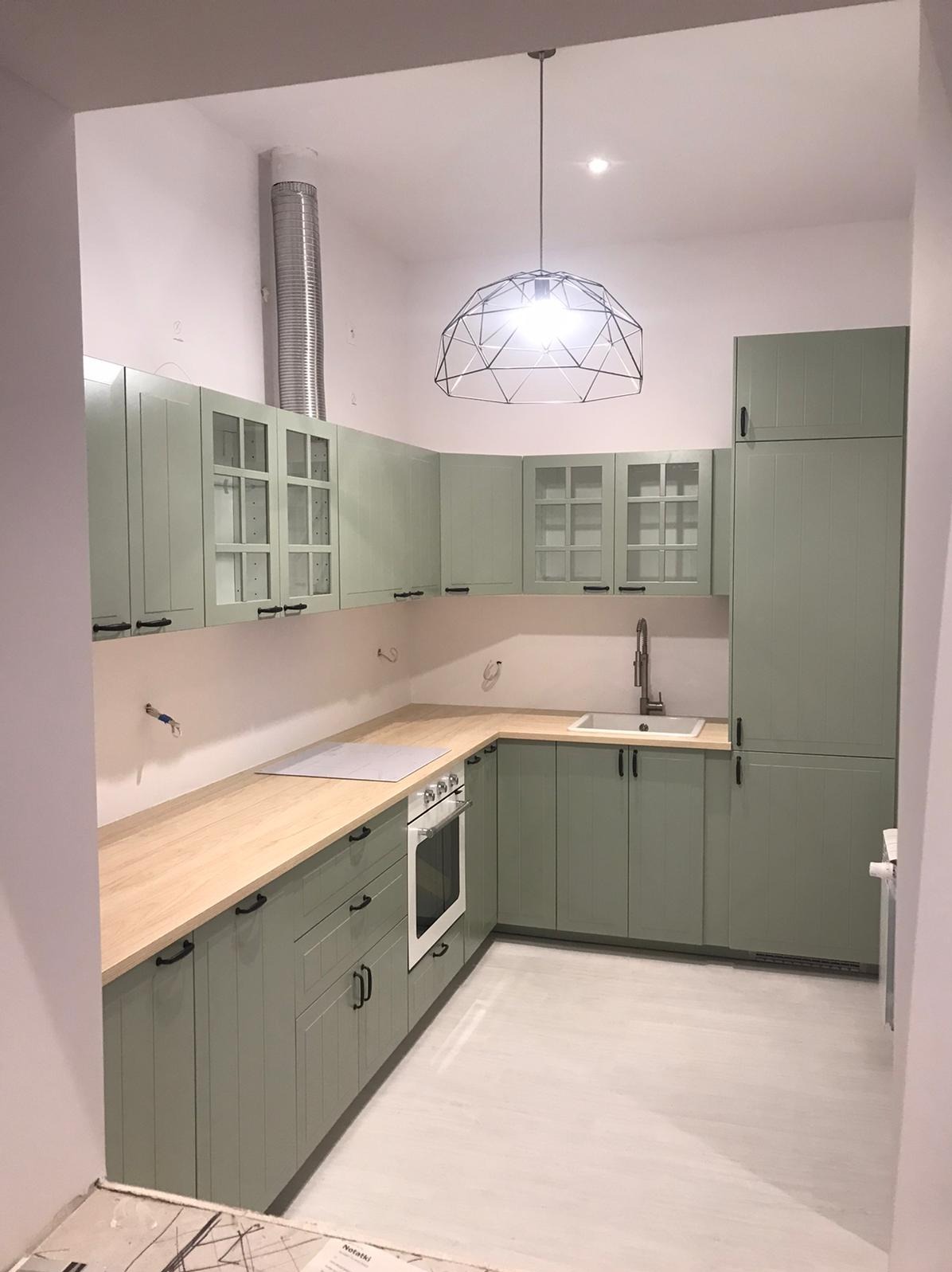 Jasnozielona kuchnia w stylu rustykalnym z drewnianym blatem, metalowym okapem i geometryczną lampą wiszącą, widok na fragment podłogi w trakcie wykańczania.