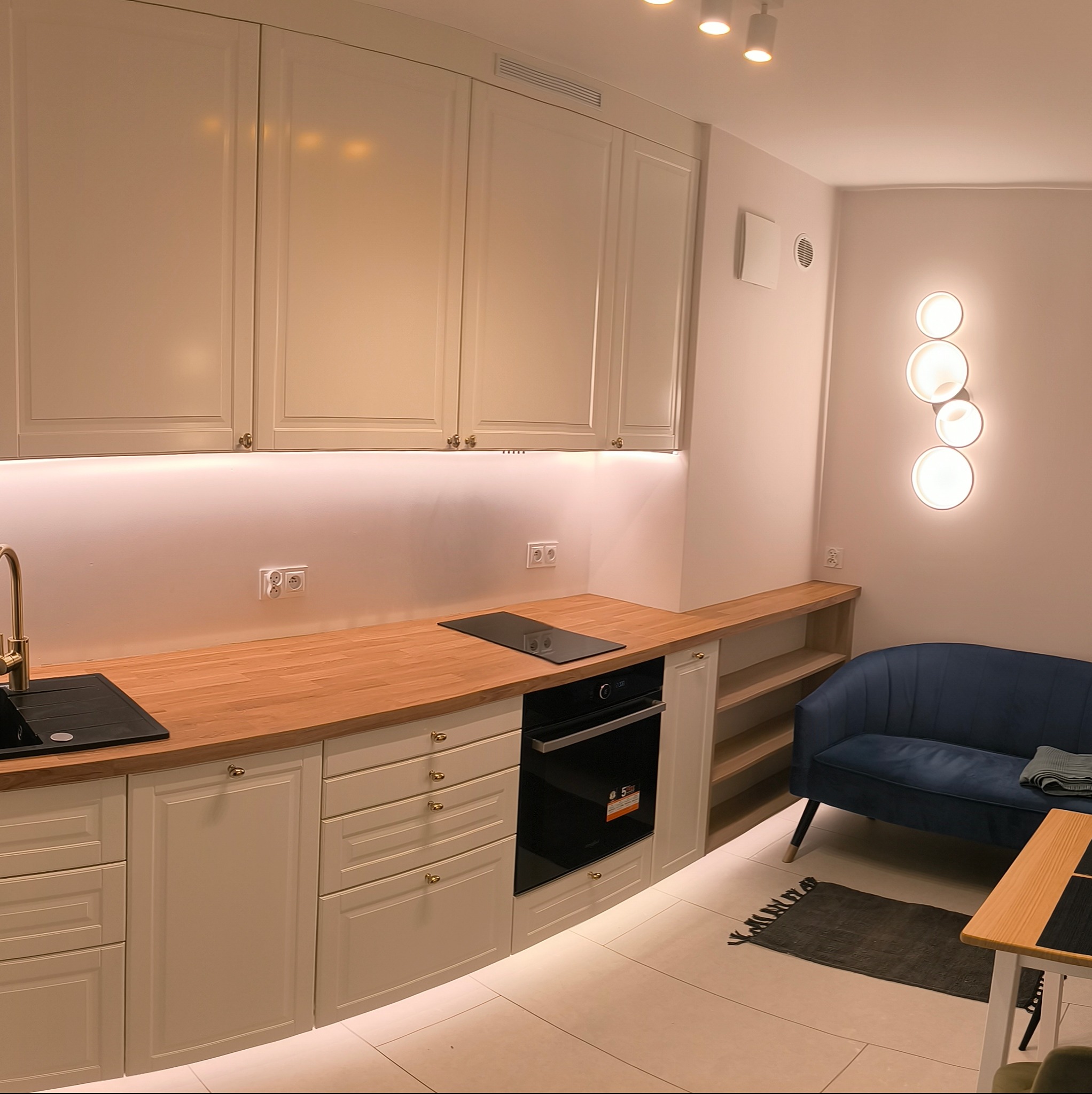 Jasna kuchnia z drewnianym blatem, oświetleniem LED pod szafkami i niebieską sofą w tle.