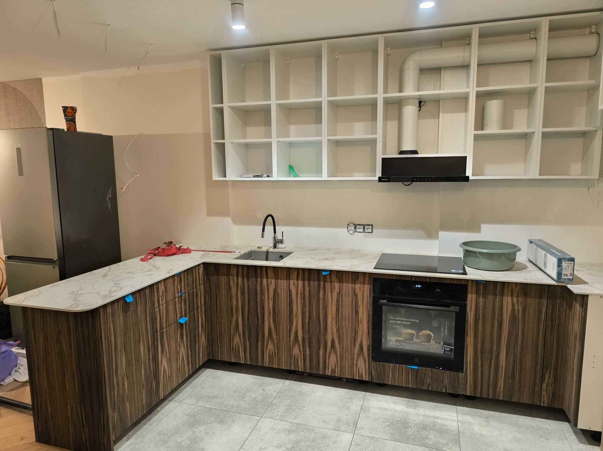 Montaż kuchni: widoczne szafki dolne z frontami w ciemnym drewnie, marmurowy blat, czarna płyta indukcyjna i piekarnik, a także białe szafki górne bez frontów i szary zlew. Na blacie miska...