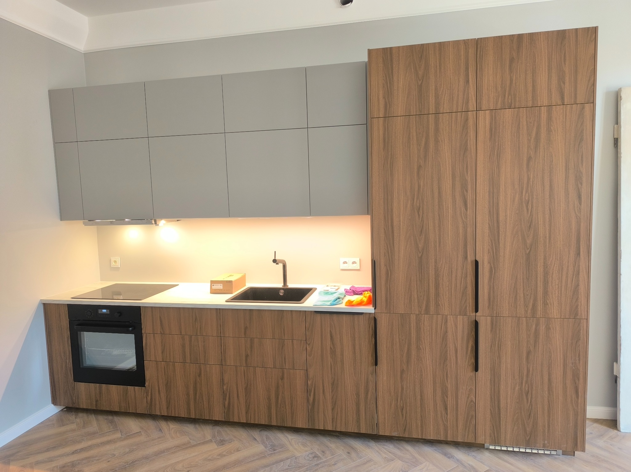 Nowoczesna kuchnia z szarymi górnymi szafkami i drewnopodobnymi dolnymi szafkami, czarnym zlewem i płytą indukcyjną, oświetlona światłem LED pod szafkami, z podłogą ułożoną w jodełkę.