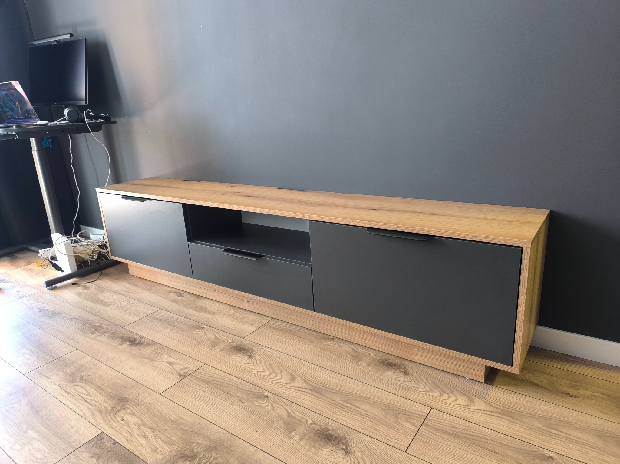 Nowoczesna szafka RTV z drewnianym blatem i grafitowymi frontami na tle szarej ściany, z widocznymi kablami i sprzętem RTV obok.