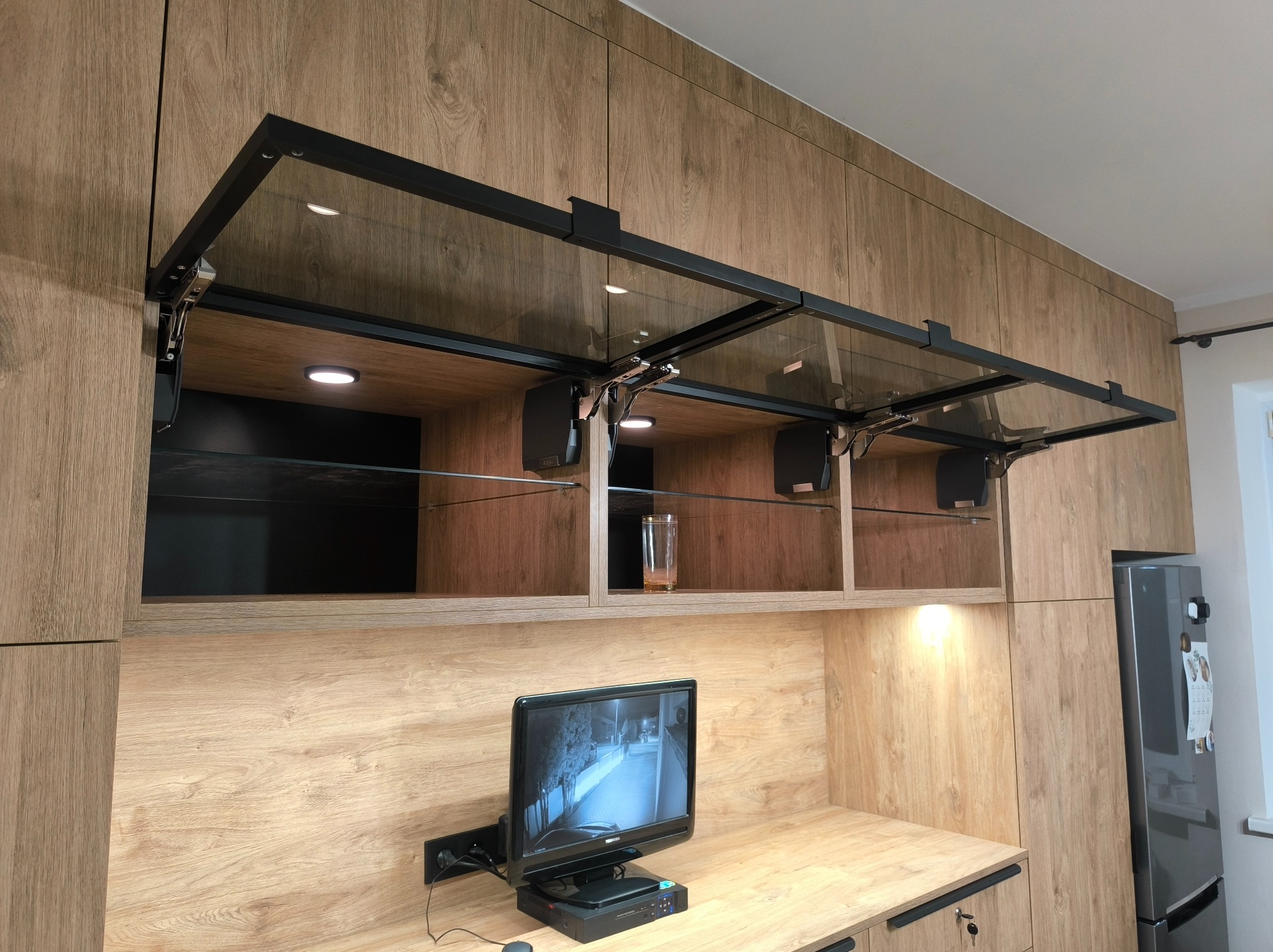 Kuchnia z drewnianymi frontami, przeszklone szafki górne z czarnymi ramami, oświetlenie punktowe, monitor na blacie.