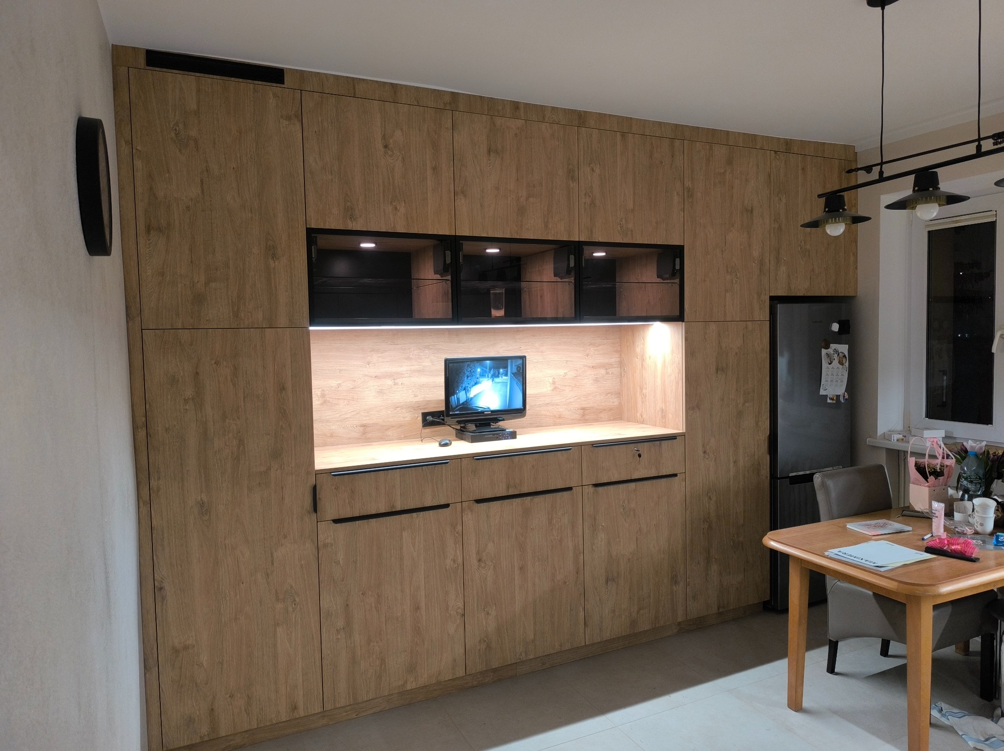 Zabudowa meblowa w salonie z drewnopodobnymi frontami, czarnymi uchwytami i wnęką z podświetleniem, w której stoi telewizor. Na ścianie wisi okrągły, czarny zegar.