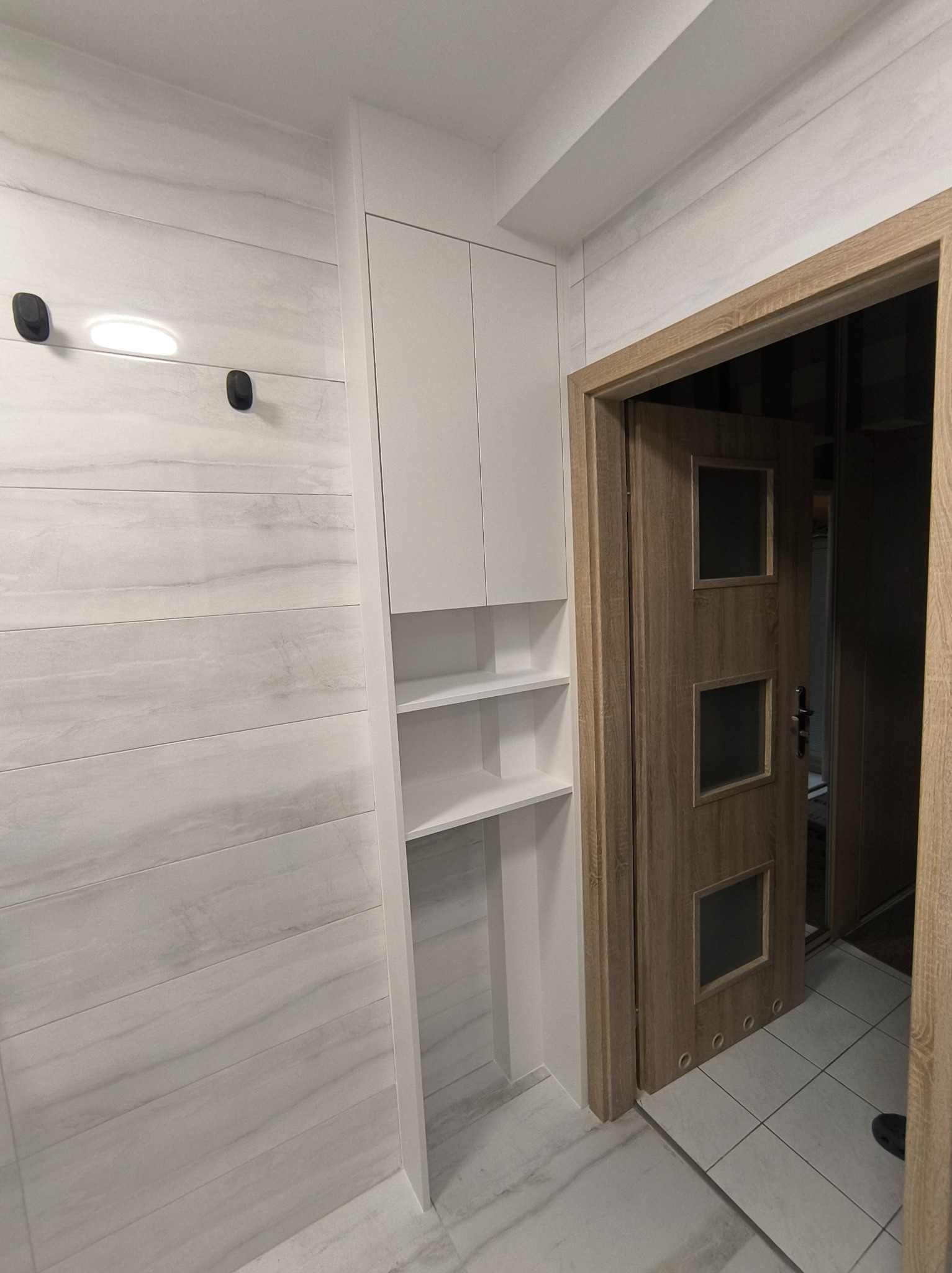 Biała szafa wnękowa z półkami, wkomponowana w przedpokój z panelami ściennymi imitującymi marmur, obok drewniane drzwi z szybkami.